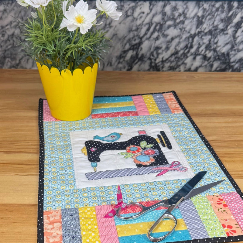 A colorful patchwork quilted table runner with a black and white sewing machine design, displayed on a wooden table next to a yellow potted plant.