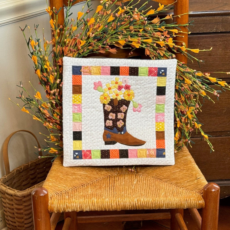 Quilted pillow featuring a cowgirl boot filled with flowers on a wooden chair with a fall flower wreath.