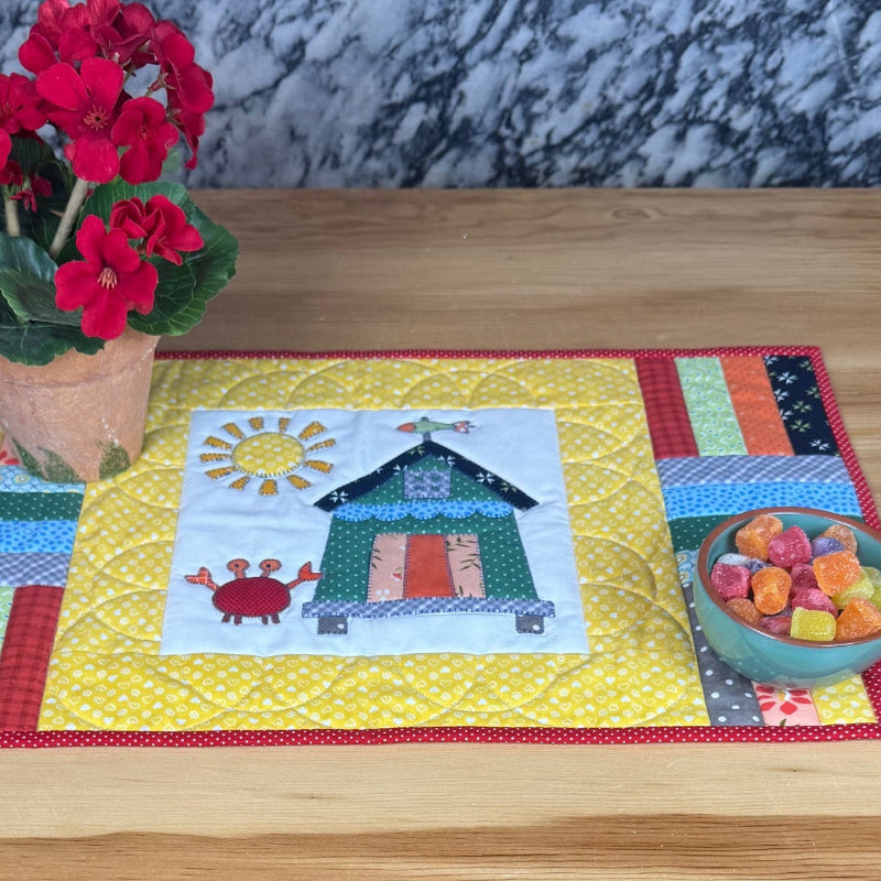 A colorful patchwork quilted placemat featuring a central cross-stitch style house design, styled on a light wood table with a potted red flower and a bowl of fruit.