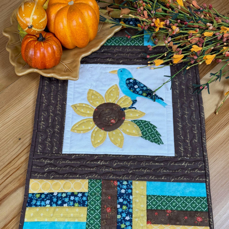 Quilted placemat featuring a blue bird on a sunflower with a leaf plate with pumpkins and some fall flowers on a wooden table.