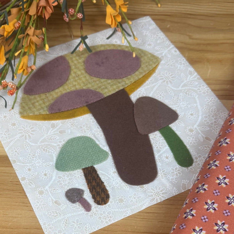 A square fabric art piece featuring a whimsical mushroom design in earthy tones, styled on a light wood tabletop next to a patterned orange fabric roll and dried orange flowers.