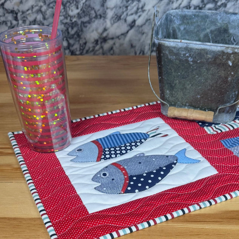 A handmade quilted patchwork placemat featuring fish designs on a red and blue patterned background, styled on a wooden table with a clear plastic tumbler and a glass container.