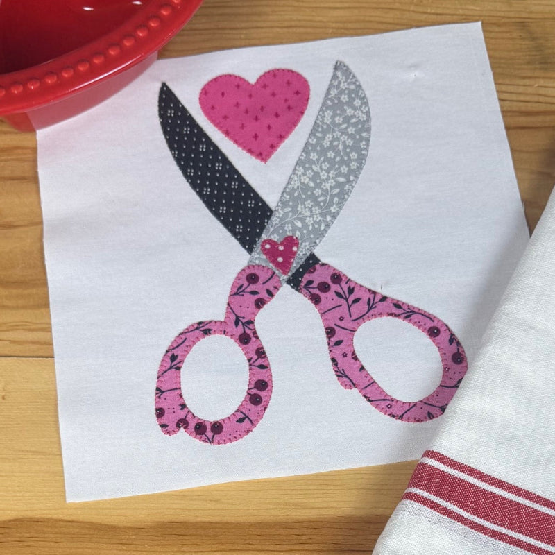 A white kitchen towel with red stripes is displayed on a wooden table next to a printed paper design of scissors and a heart, with a red heart-shaped dish visible in the top left.