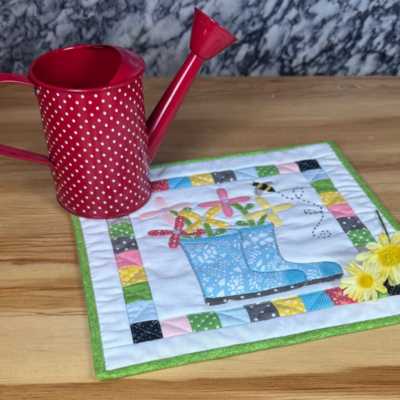 A small square quilted fabric placemat with a colorful patchwork border and a blue watering can design sits on a light wood table next to a red polka-dot watering can.