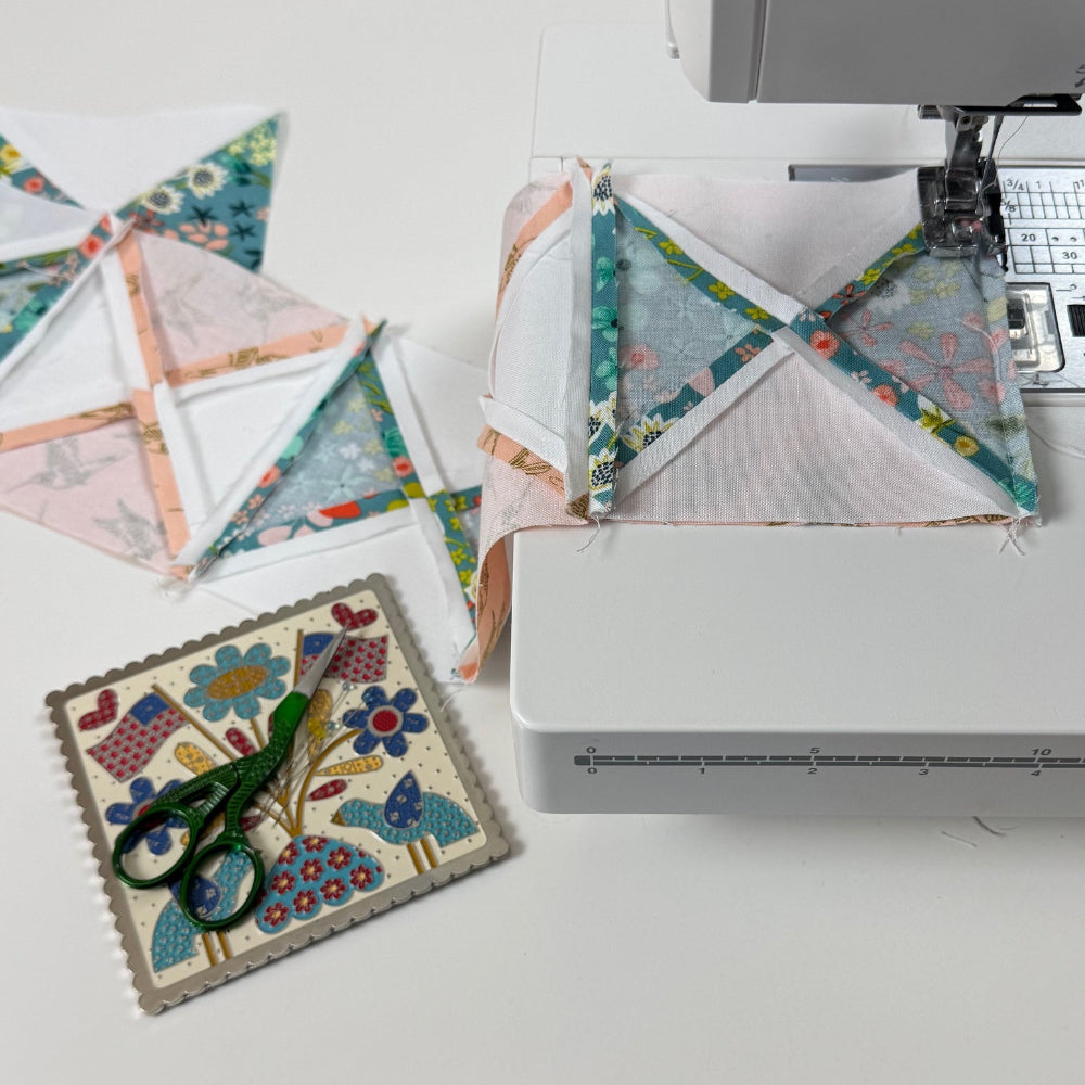 Quilt blocks being sewn by a sewing machine on a white surface with stork scissors on Americana themed square.