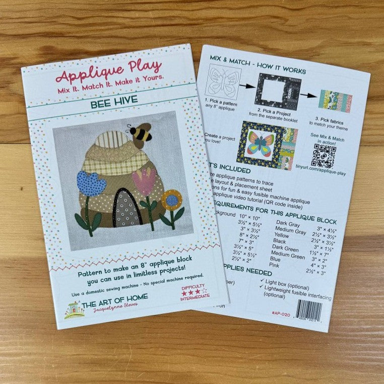 Front and back covers of an applique block pattern titled "Applique Play" by The Art of the Home, placed on a wooden surface. One sheet shows the front cover with a bee and flower applique design, and the other shows instructions and included colors.