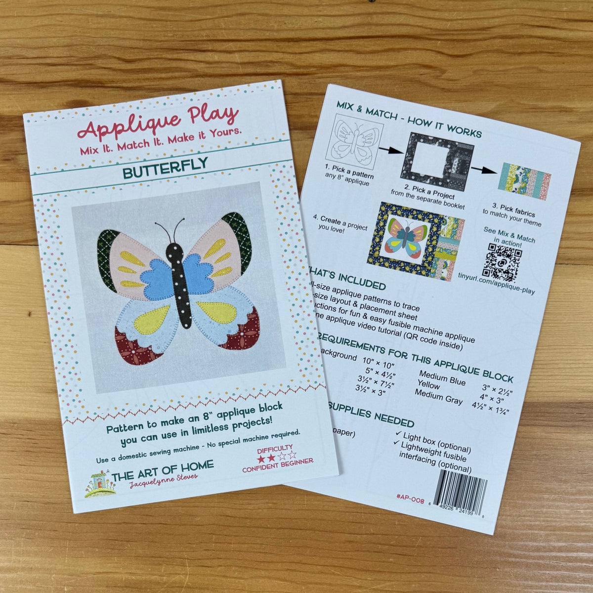 Front and back covers for an applique block pattern titled "Butterfly" by The Art of the Home, placed on a wooden surface. One sheet shows the front with a butterfly design and instructions, and the other shows the back with included supplies and a QR code.