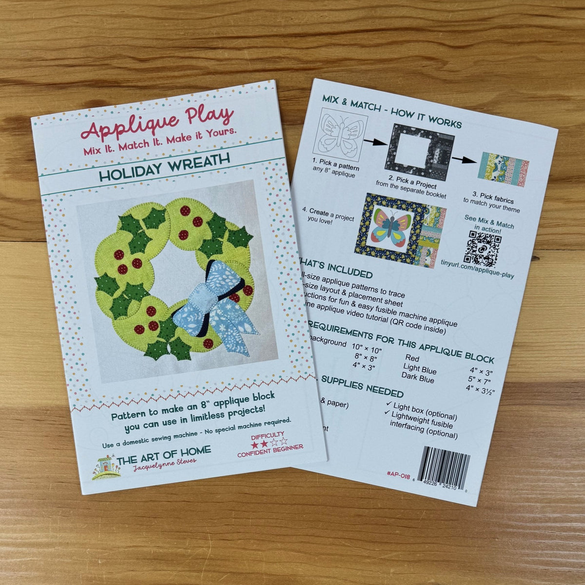 Front and back covers for 'Applique Play' Holiday Wreath patterns laid on a wooden surface. One card shows the front cover with a wreath illustration and product title, and the other shows an instruction panel with step-by-step guidance and included supplies.