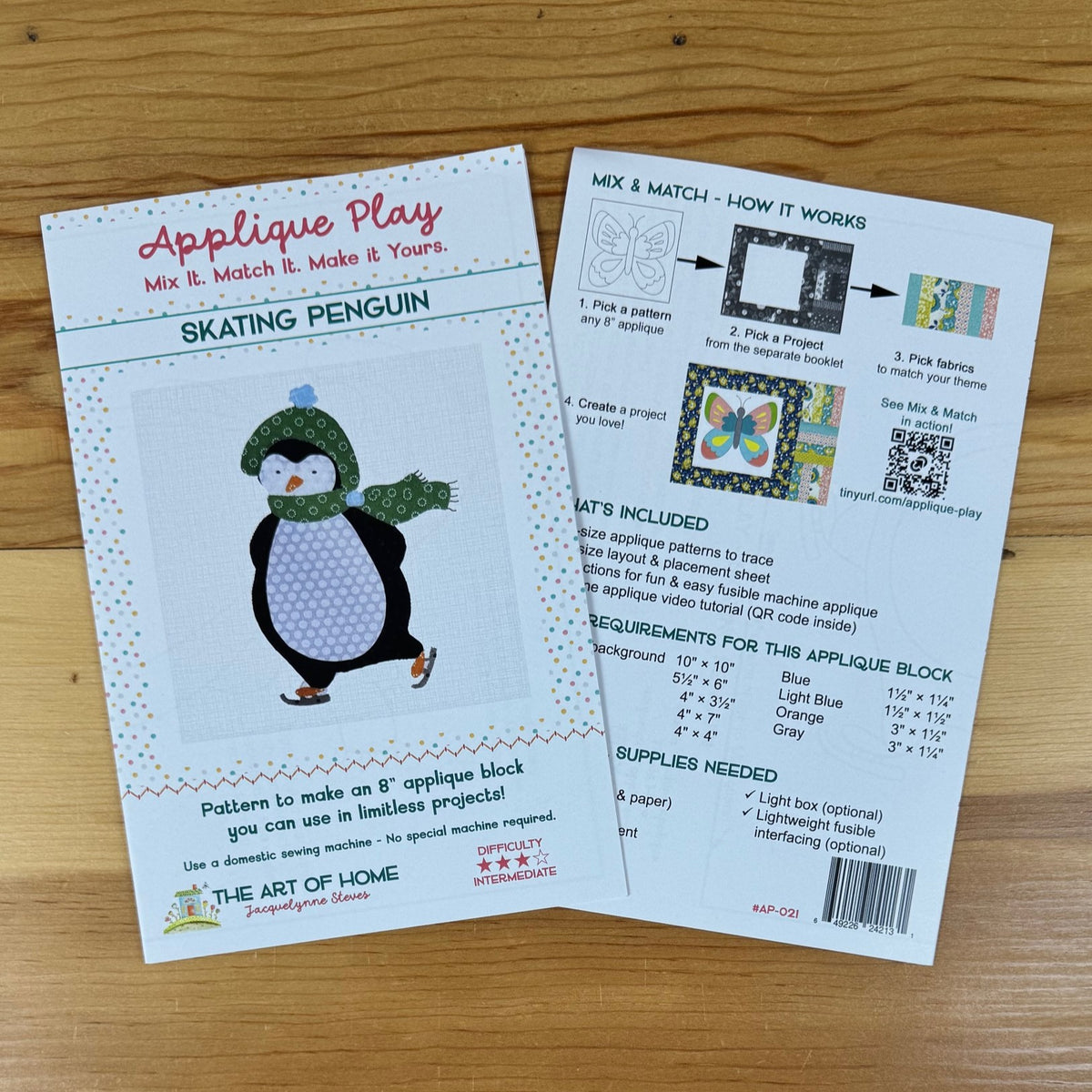 Front and back covers of pattern for making a Skating Penguin applique block, placed on a light wood surface. One sheet shows the front cover with a penguin illustration and title text, and the other shows the back with instructions, included materials, and a QR code.