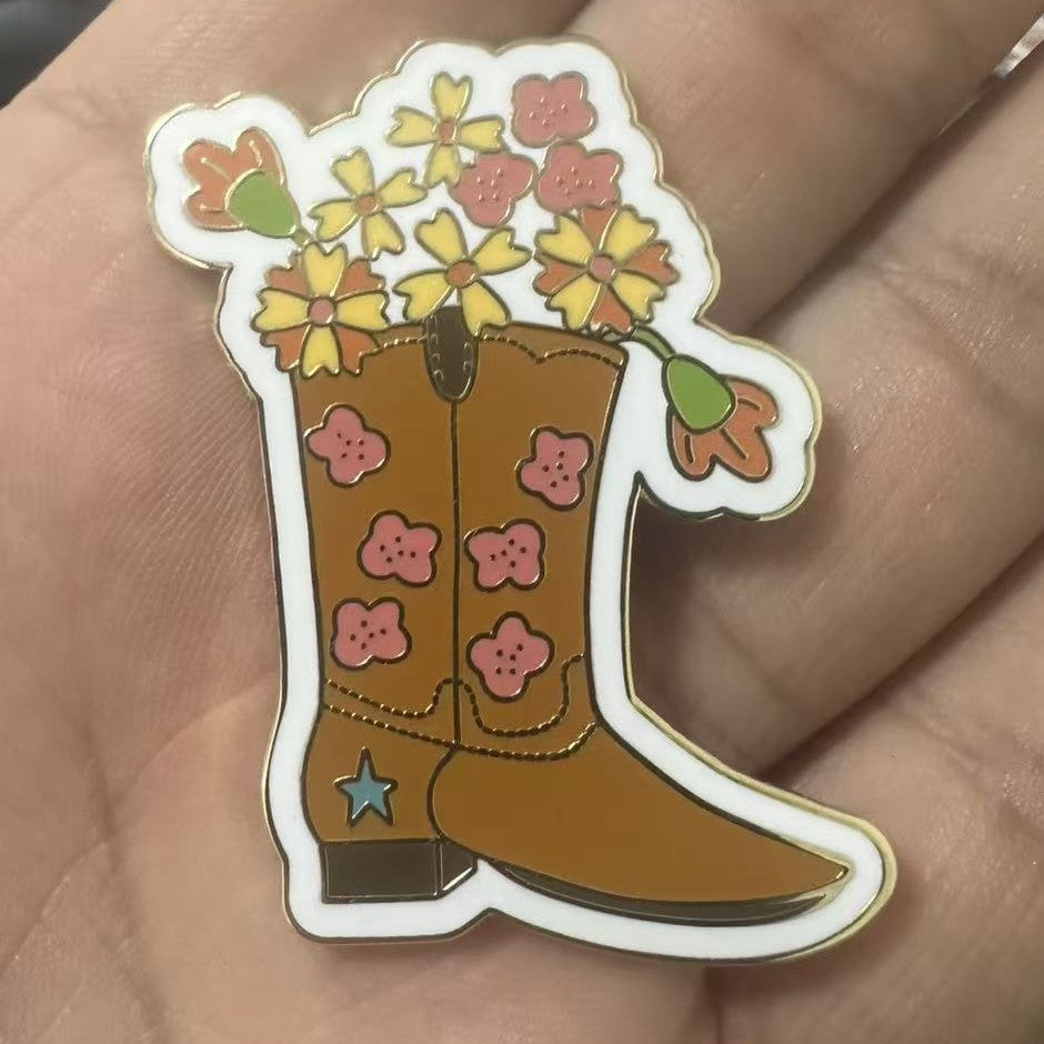 A cowboy boot enamel needle minder with pink flowers and yellow flowers growing out of the boot, held in a person's hand.