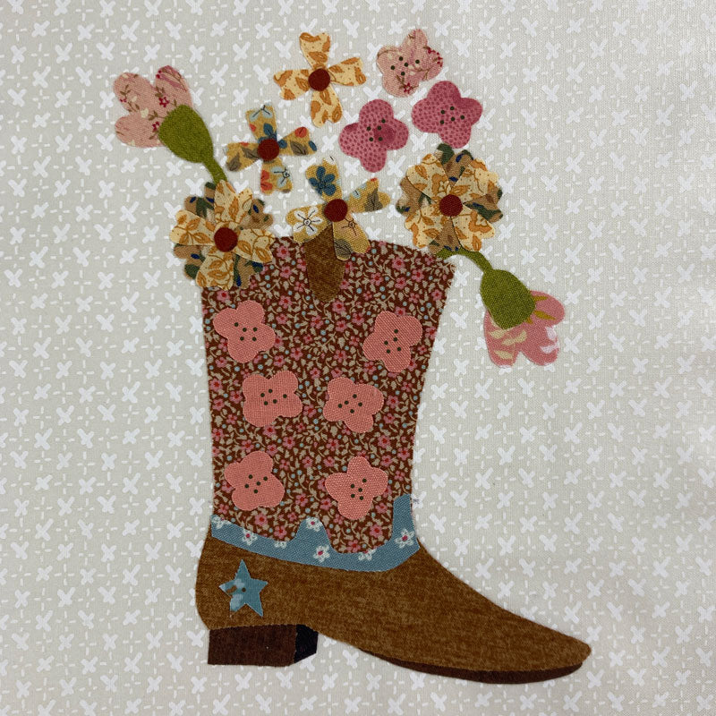 A quilted fabric applique of a brown cowboy boot filled with a bouquet of colorful floral patches, set against a light beige patterned fabric background.
