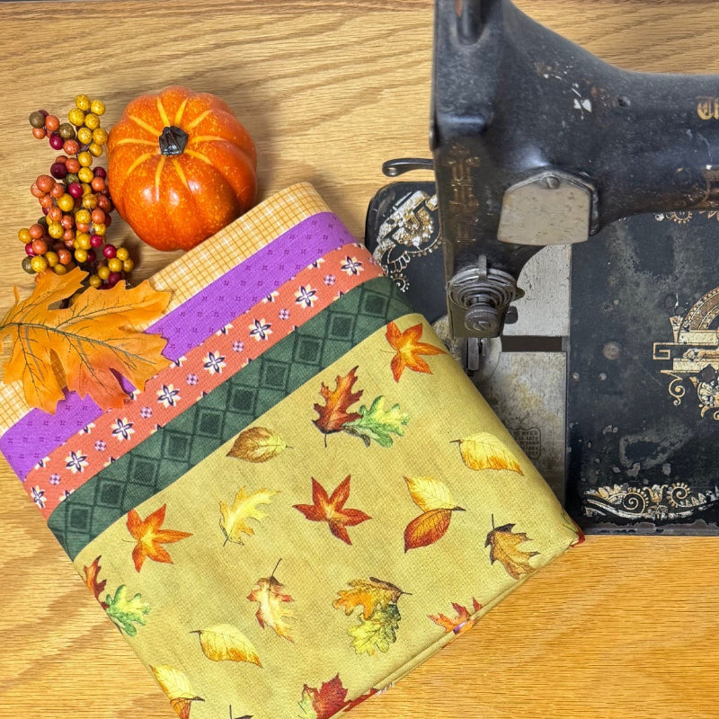 A folded piece of yellow fabric with an autumn leaf pattern and striped border is displayed on a wooden table next to a vintage sewing machine.