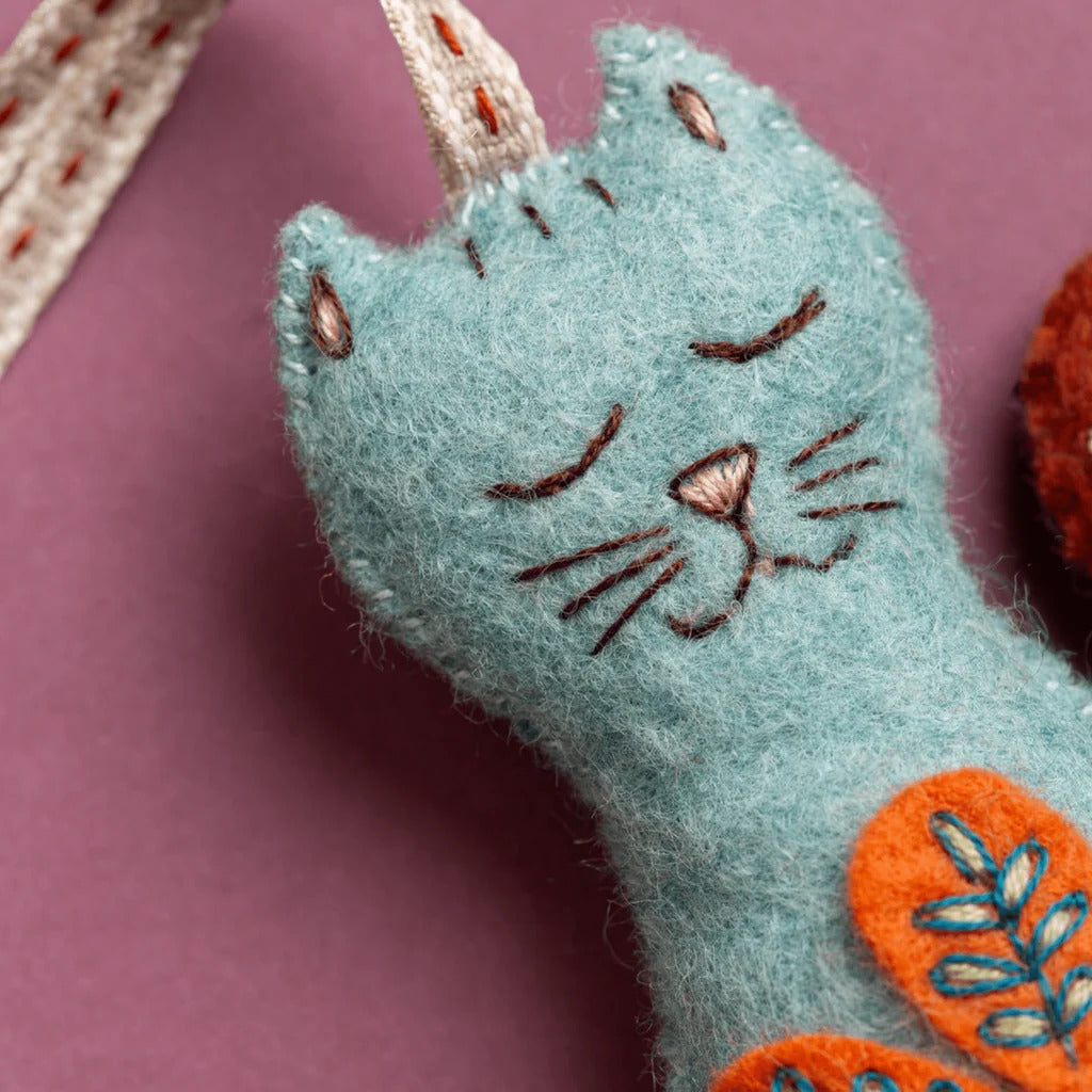 Blue cat-shaped felt ornament with orange folk embroidered designs on a pink background