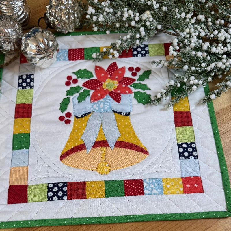 A square quilted fabric placemat or table topper featuring a festive Christmas design with two yellow bells, a red poinsettia, green holly leaves, and a multicolor patchwork border, displayed on a wooden tabletop with holiday greenery and silver ornaments.