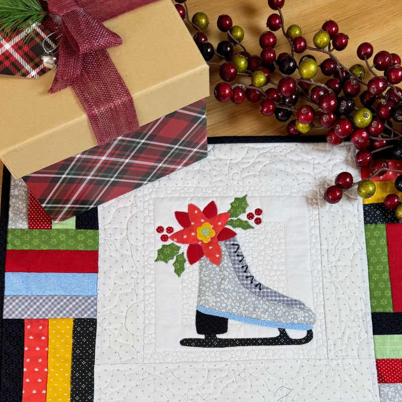 A square quilted fabric placemat or runner featuring a stitched gray ice skate with a red poinsettia and green leaves on a white center panel, surrounded by multicolored patchwork borders. It is displayed on a wooden table with holiday decorations, including a wrapped gift box and red berry branches.