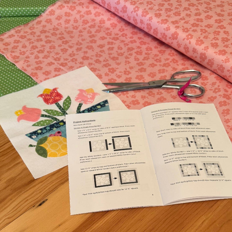 A tabletop display of a quilting or fabric crafting kit, featuring a pink patterned fabric roll, a green patterned fabric roll, an open instruction booklet with quilting templates, and a floral patterned paper sheet, all arranged on a wooden table.