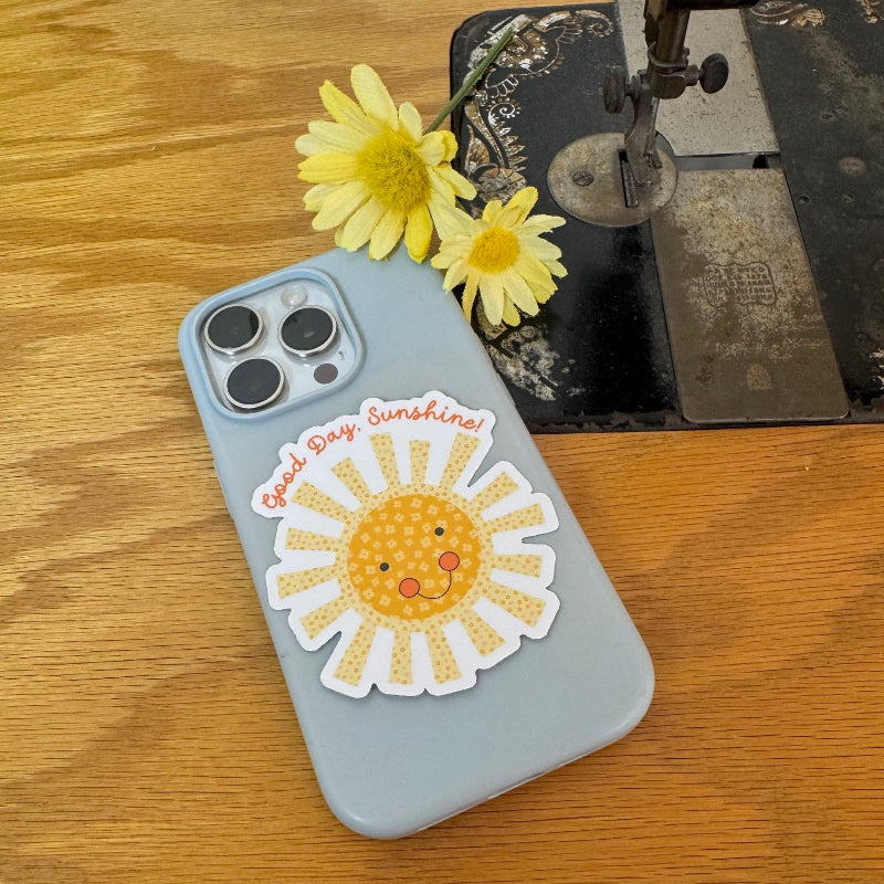 A light blue phone case with a smiling yellow sun sticker and the words 'Good Day Sunshine!' in red script, resting on a wooden table next to yellow daisies and a vintage sewing machine.
