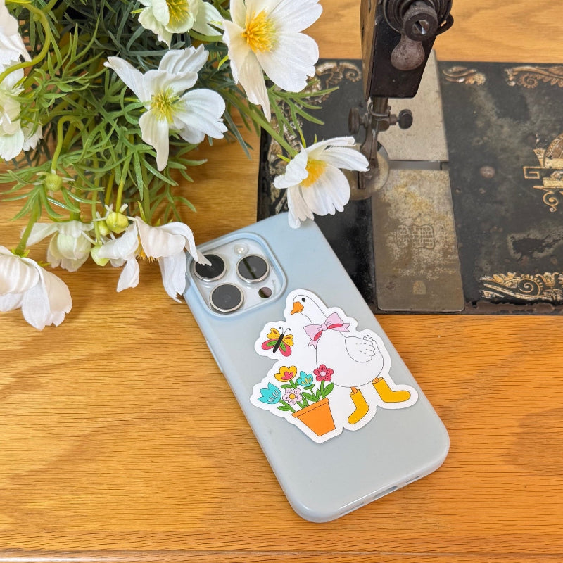 A light blue phone case with a colorful duck and flowers sticker, resting on a wooden table next to white daisies and a vintage sewing machine.