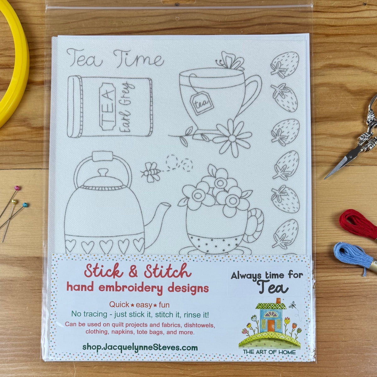 A clear plastic sleeve containing a white fabric design sheet with black line drawings of tea-themed motifs, including a teapot, teacup, flowers, tea bag, and strawberries, plus printed product text. The sheet is photographed on a wooden surface with sewing supplies like pins, scissors, thread, and ribbon around it.