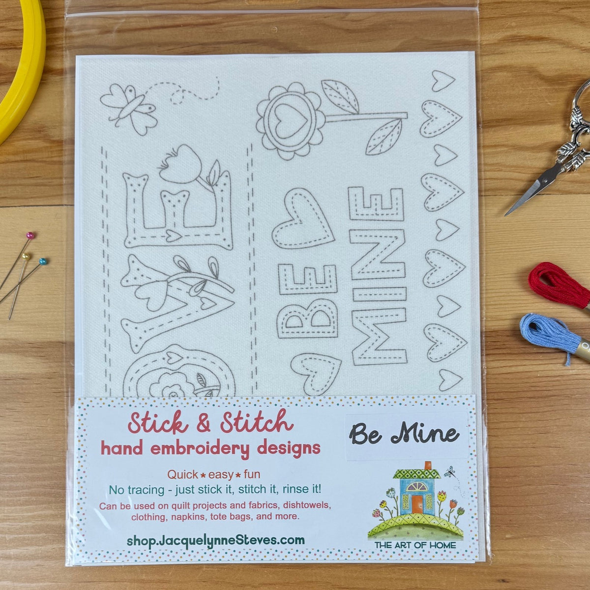 A packaged stick and stitch hand embroidery design kit featuring valentine themed 'Be Mine' designs, displayed on a wooden tabletop with embroidery floss, pins, and scissors nearby.