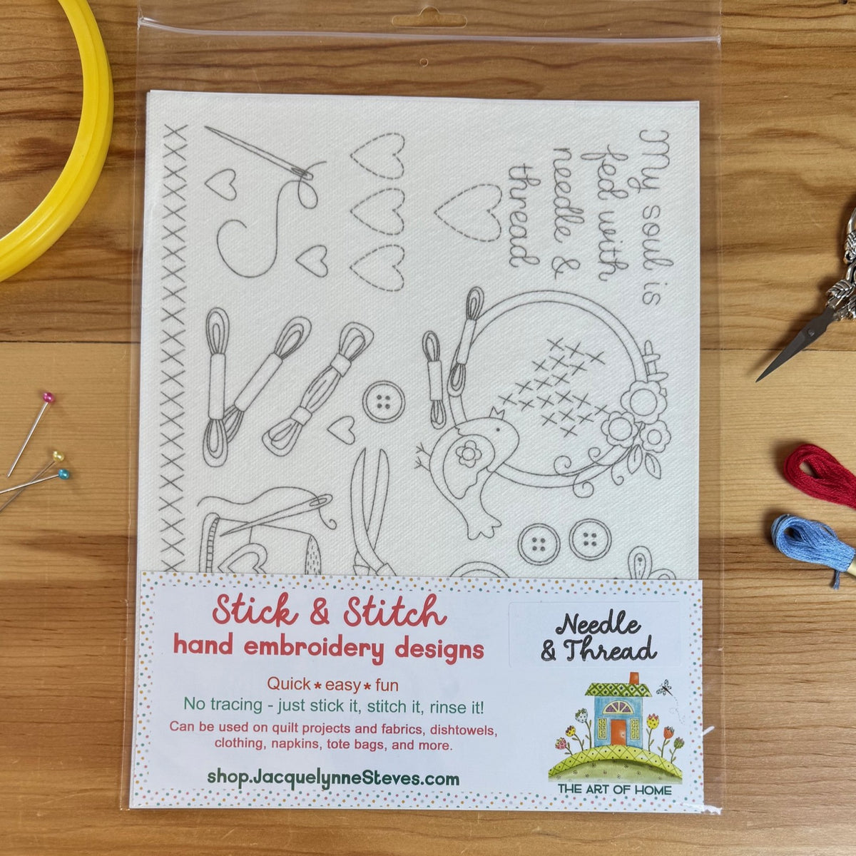 A clear plastic bag containing a hand embroidery design kit sheet labeled 'Stick & Stitch hand embroidery designs' and 'Needle & Thread' by The Art of Home, placed on a wooden surface with sewing tools around it.
