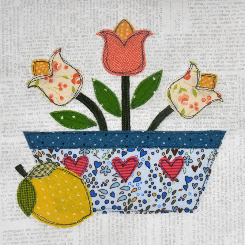 A handmade fabric applique design depicting three tulips in a patterned blue and white pot, with a yellow lemon in the foreground, set against a background of vintage book pages.