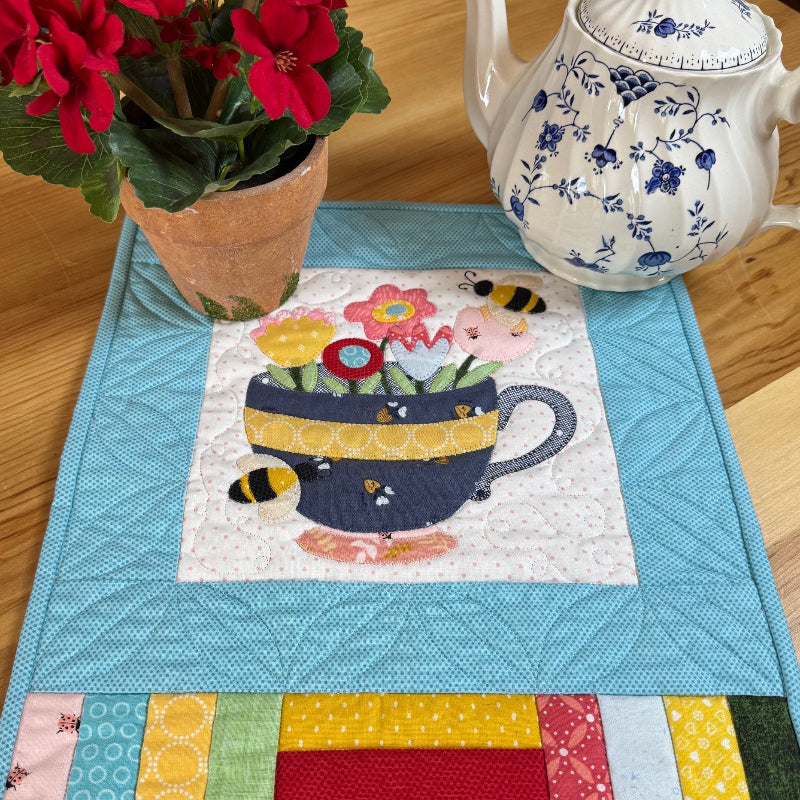 A quilted fabric table runner with a colorful patchwork border and a central floral teacup design, styled on a wooden table next to a potted red flower and a blue-and-white teapot.