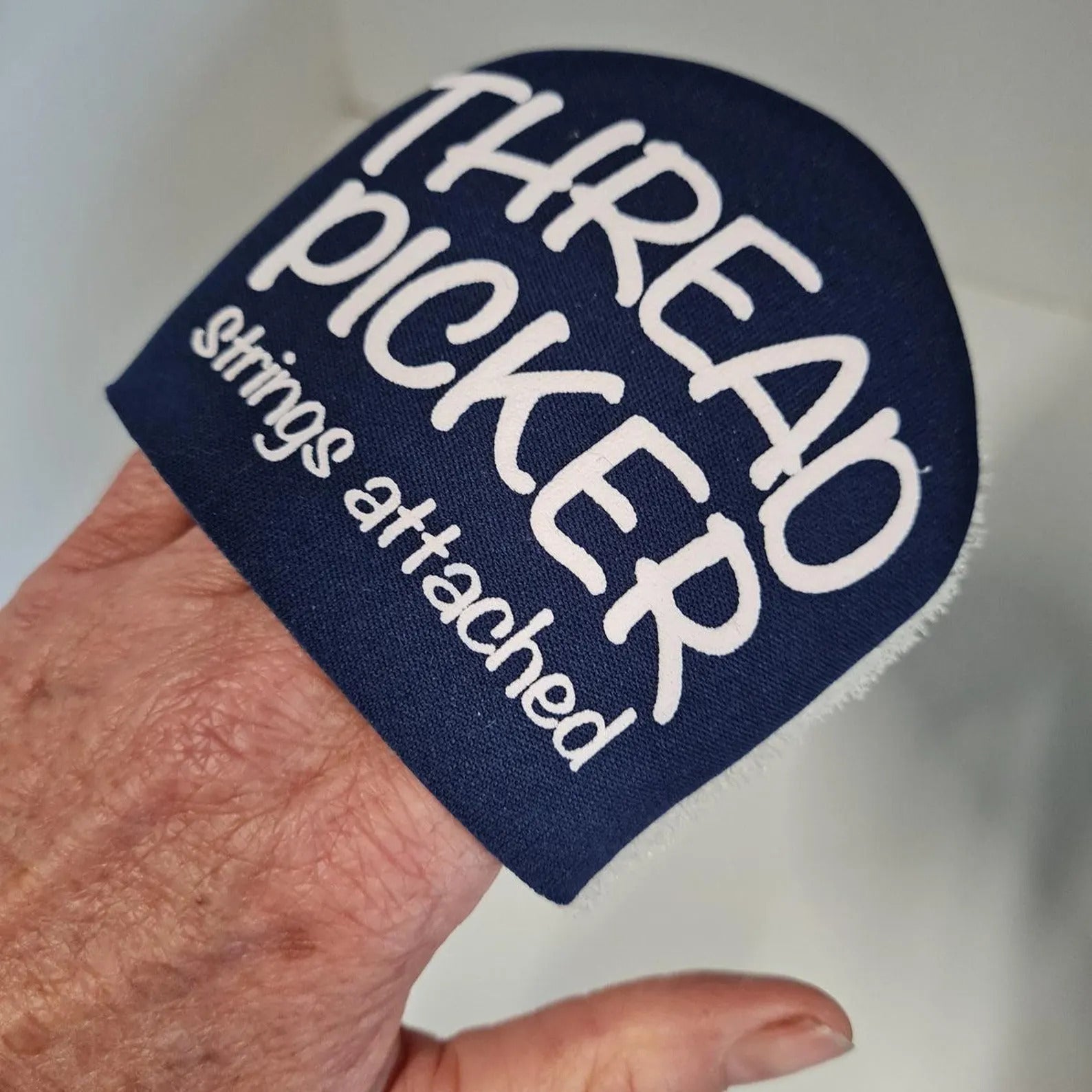 Navy blue patch with white text 'THREAD PICKER strings attached' held by a hand against a white background