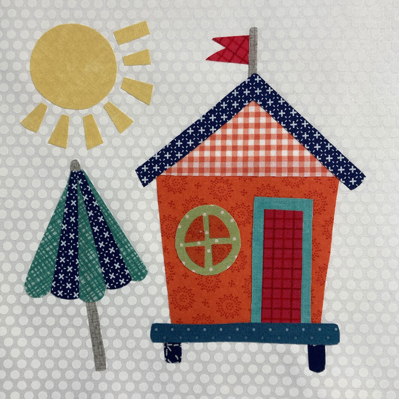 A whimsical applique design of a small house with a red-and-white gingham roof, orange body, teal trim, a red door, and a green window. A yellow sun and a teal-and-navy umbrella appear on a light gray polka-dot background.