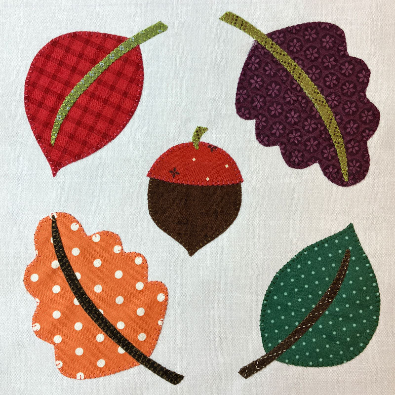 A square with applique design featuring five autumn-themed applique shapes: two leaves, an acorn, and two additional leaves, each with different patterns like plaid, polka dots, and floral prints on a light beige linen background.
