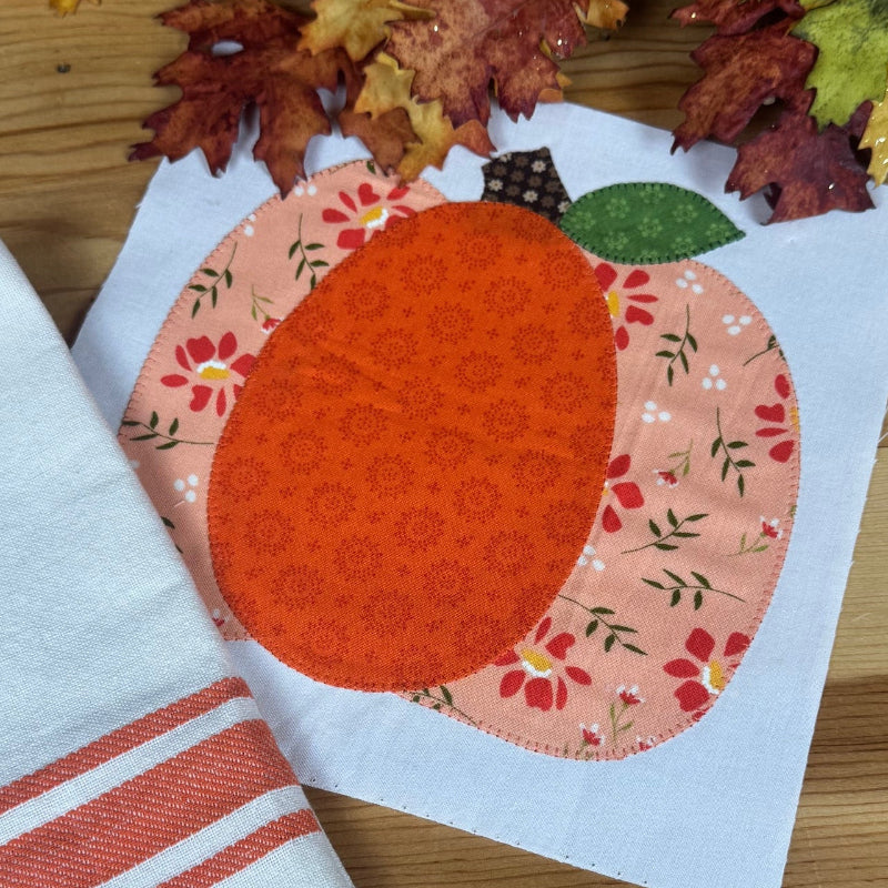 A folded white kitchen towel with two orange stripes lies on a wooden table next to a white napkin featuring a decorative orange pumpkin applique. Artificial autumn leaves are scattered at the top of the frame.