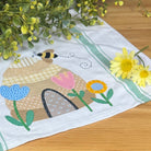 A white kitchen tea towel with a colorful embroidered bee and flower design, styled on a wooden table with yellow flowers.