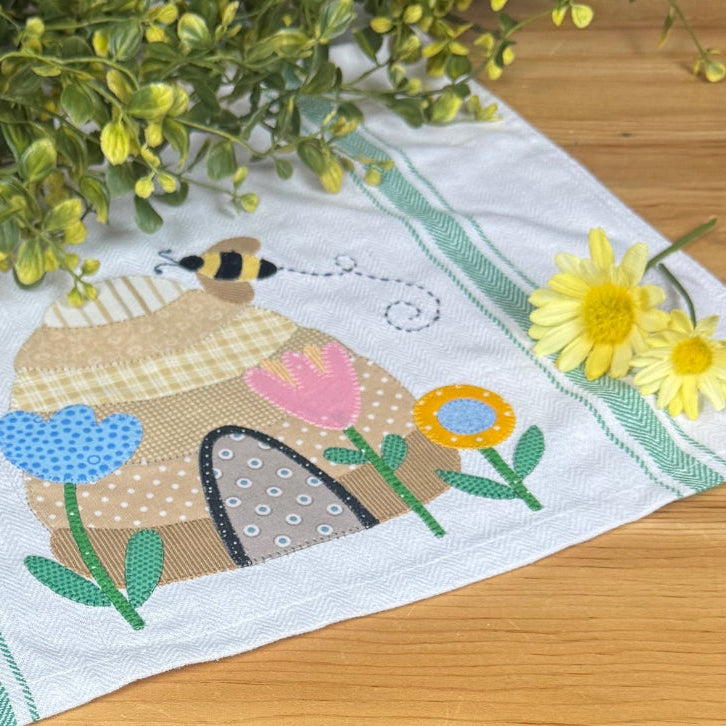 A white kitchen tea towel with a colorful embroidered bee and flower design, styled on a wooden table with yellow flowers.