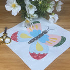 A square of white fabric featuring a colorful butterfly applique design, laid on a wooden table with gold-handled scissors and a small bouquet of white flowers nearby.