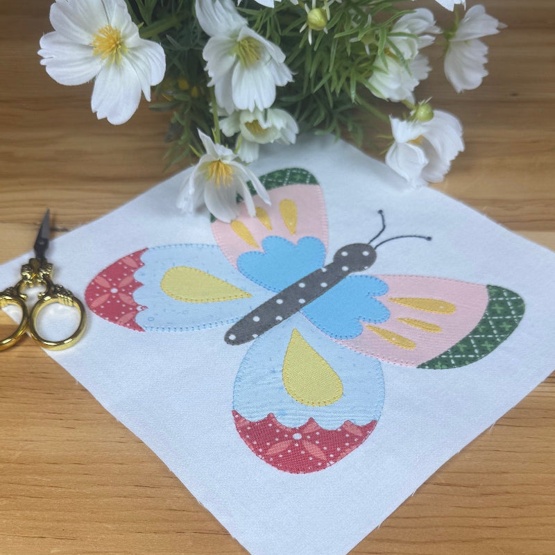 A square of white fabric featuring a colorful butterfly applique design, laid on a wooden table with gold-handled scissors and a small bouquet of white flowers nearby.