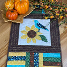 Quilted placemat featuring a blue bird on a sunflower with a leaf plate with pumpkins and some fall flowers on a wooden table.