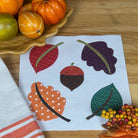 A styled autumn flat-lay featuring a white kitchen towel with coral stripes, a white paper napkin with colorful embroidered leaves and acorns, decorative fall leaves, berries, and small pumpkins on a wooden table.