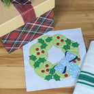 A holiday-themed gift set on a light wood table, featuring a kraft paper gift box wrapped in a burgundy ribbon with a small pine sprig, a stack of plaid gift boxes, a printed Christmas wreath card, and a folded white cloth napkin with green stripes.
