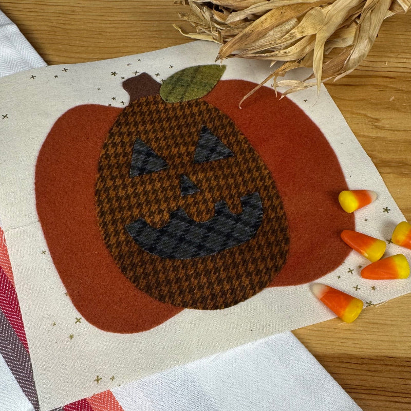 A square cream fabric napkin or small table linen laid on a wooden surface, featuring a stitched jack-o'-lantern design in rust orange, dark brown houndstooth, and gray. The napkin is styled with candy corn and dried corn husks, alongside a white striped textile.