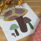 A square fabric art piece featuring a whimsical mushroom design in earthy tones, styled on a light wood tabletop next to a patterned orange fabric roll and dried orange flowers.