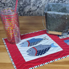 A handmade quilted patchwork placemat featuring fish designs on a red and blue patterned background, styled on a wooden table with a clear plastic tumbler and a glass container.