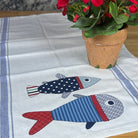 A white cotton towel with blue stripes and two embroidered fish motifs in patriotic red, white, and blue, displayed on a wooden table next to a potted red flower.