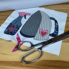 A pair of silver scissors resting on a white fabric patch featuring a patchwork design of a spray bottle, a gingham iron, and two pink hearts, all placed on a light wooden table.