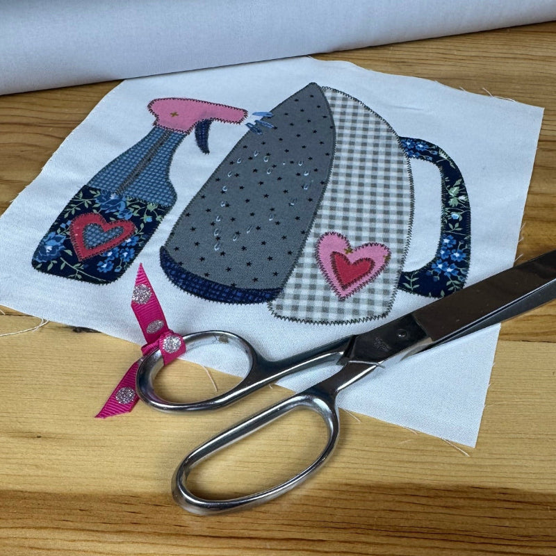 A pair of silver scissors resting on a white fabric patch featuring a patchwork design of a spray bottle, a gingham iron, and two pink hearts, all placed on a light wooden table.