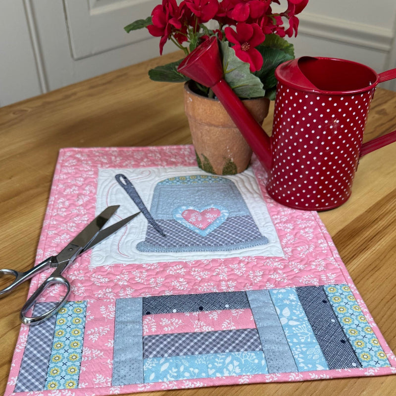 Quilted placemat featuring a thimble and needle with sewing scissors, flower pot and watering can on a wood table.