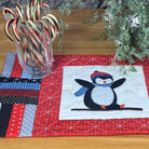 A handmade quilted table runner with a penguin design, displayed on a wooden table with holiday decorations including candy canes and a small wreath.