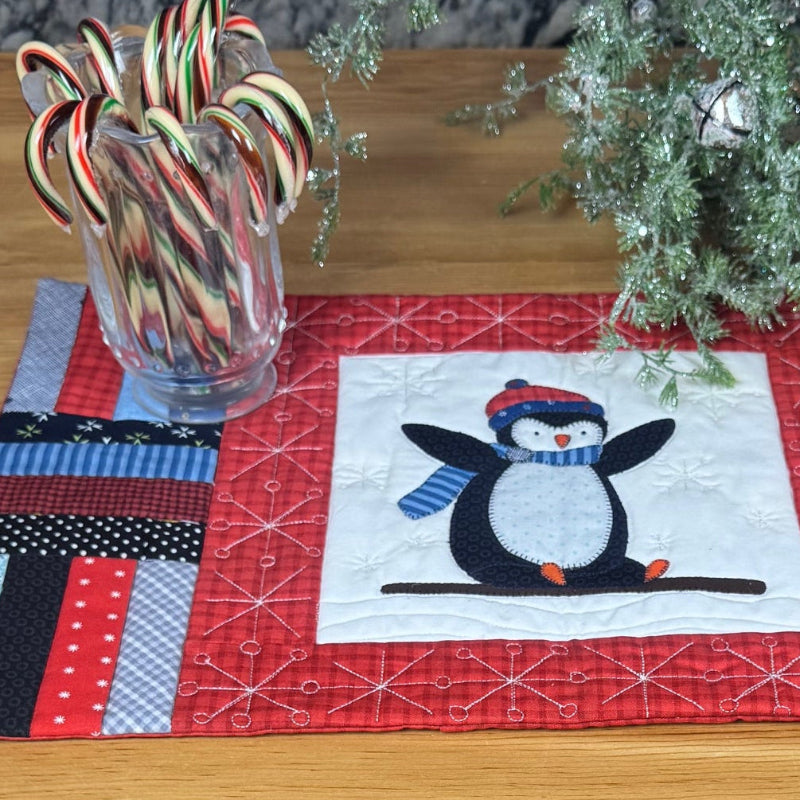 A handmade quilted table runner with a penguin design, displayed on a wooden table with holiday decorations including candy canes and a small wreath.