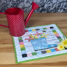 A small square quilted fabric placemat with a colorful patchwork border and a blue watering can design sits on a light wood table next to a red polka-dot watering can.