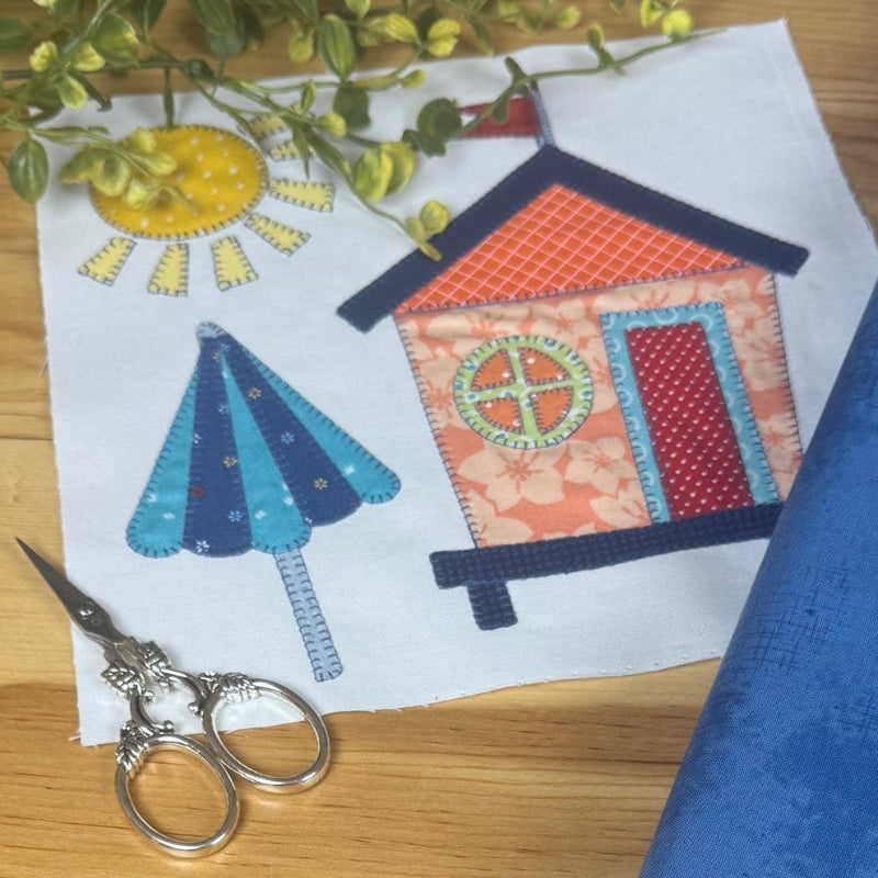A blue fabric  lies on a wooden table beside a small printed pattern card with a house and tree design, along with scissors and greenery.