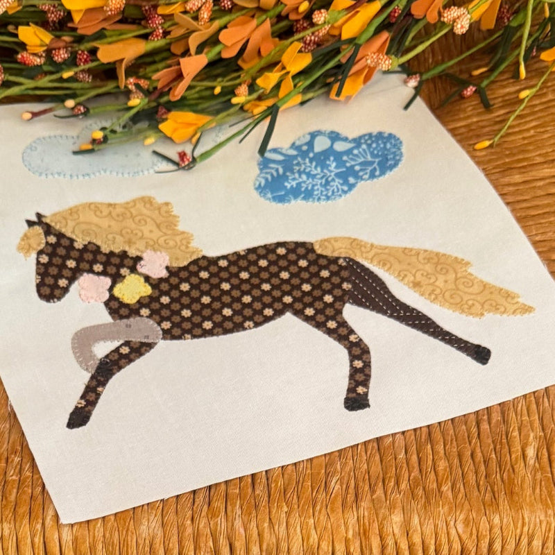 A square white fabric featuring a whimsical embroidered design of a brown horse with a gold mane and a blue cloud, displayed on a woven rattan chair.